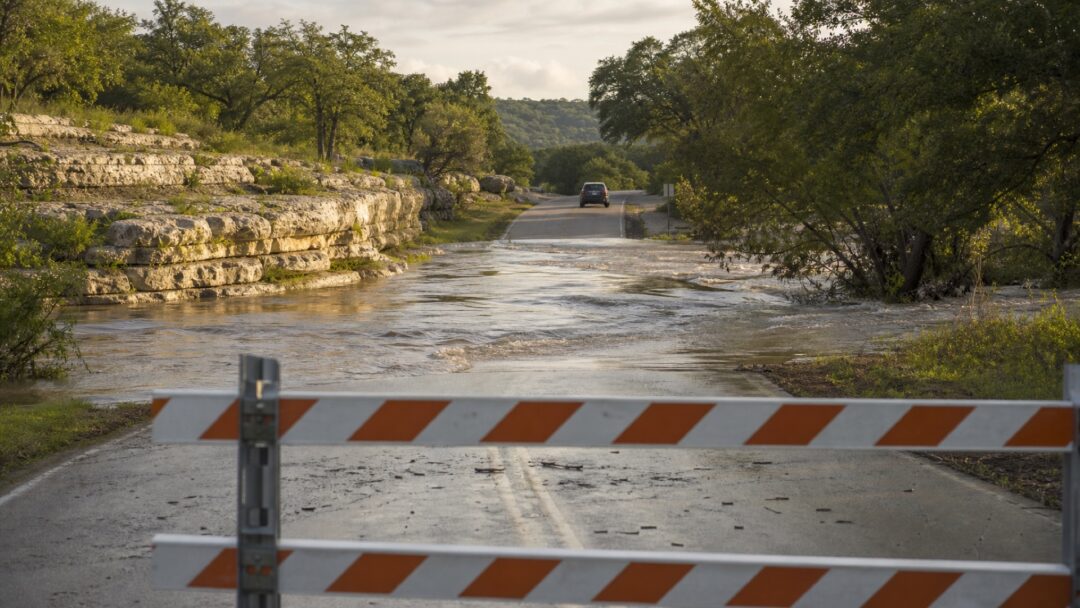 A vehicle safely turning away from moving floodwater at a barricaded Texas Hill Country low-water crossing.