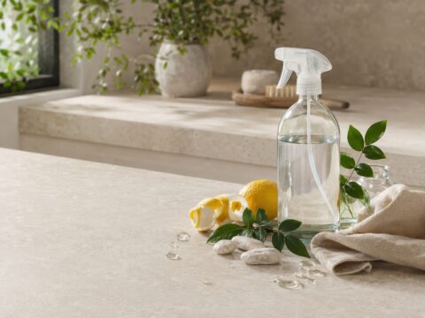 Clear refillable spray bottle with citrus, green leaves, stones, and linen on a sunlit travertine kitchen counter.