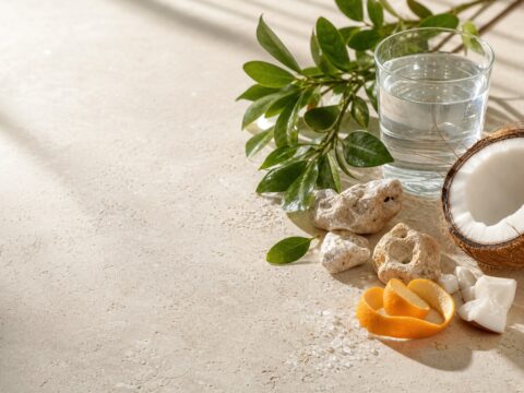 Coconut, citrus, green leaves, mineral stones, water, and clean neutral materials arranged on pale travertine.