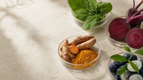 Green leaves, beet, turmeric, and deep blue botanical materials arranged in clear glass dishes on travertine.