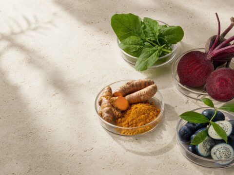 Green leaves, beet, turmeric, and deep blue botanical materials arranged in clear glass dishes on travertine.