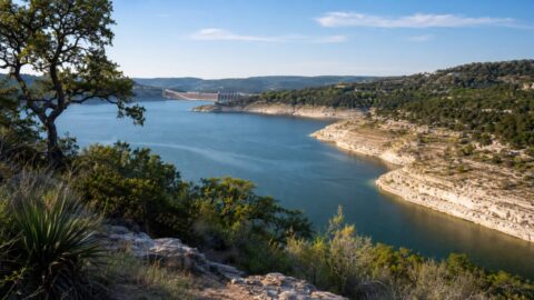 Lake Travis with exposed limestone shelves, blue-green water, live oaks, and reservoir infrastructure in the distance.