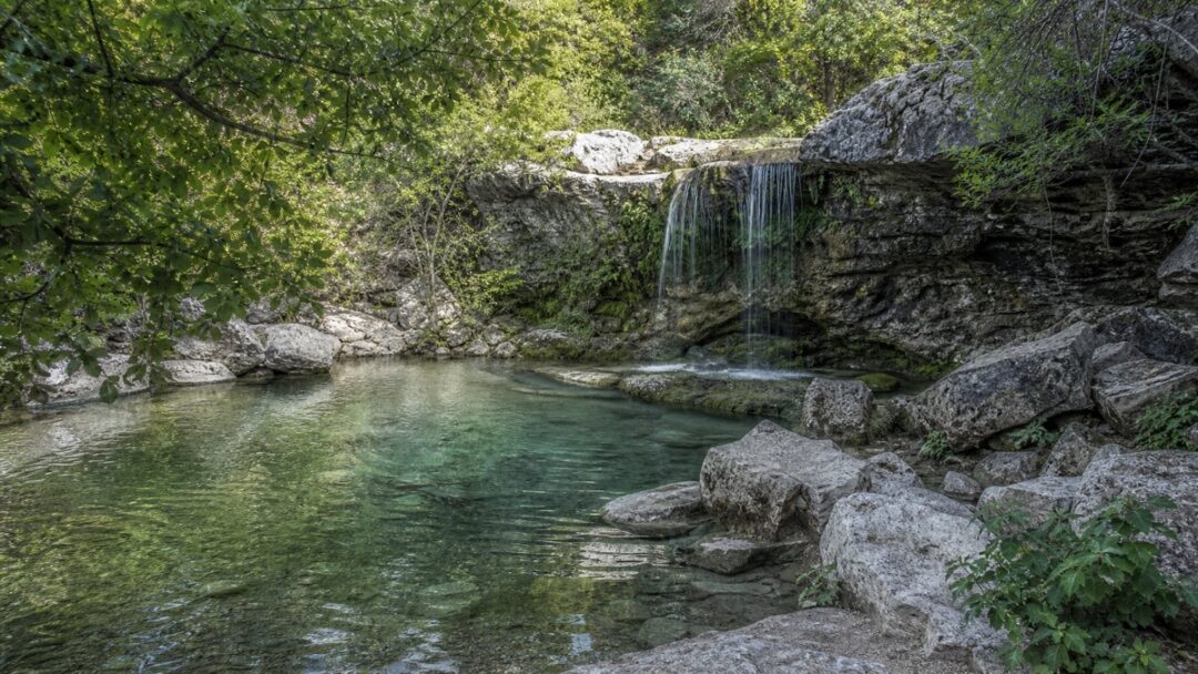 A limestone grotto and natural pool beneath a small waterfall, surrounded by shaded native canyon vegetation in the Texas Hill Country.
