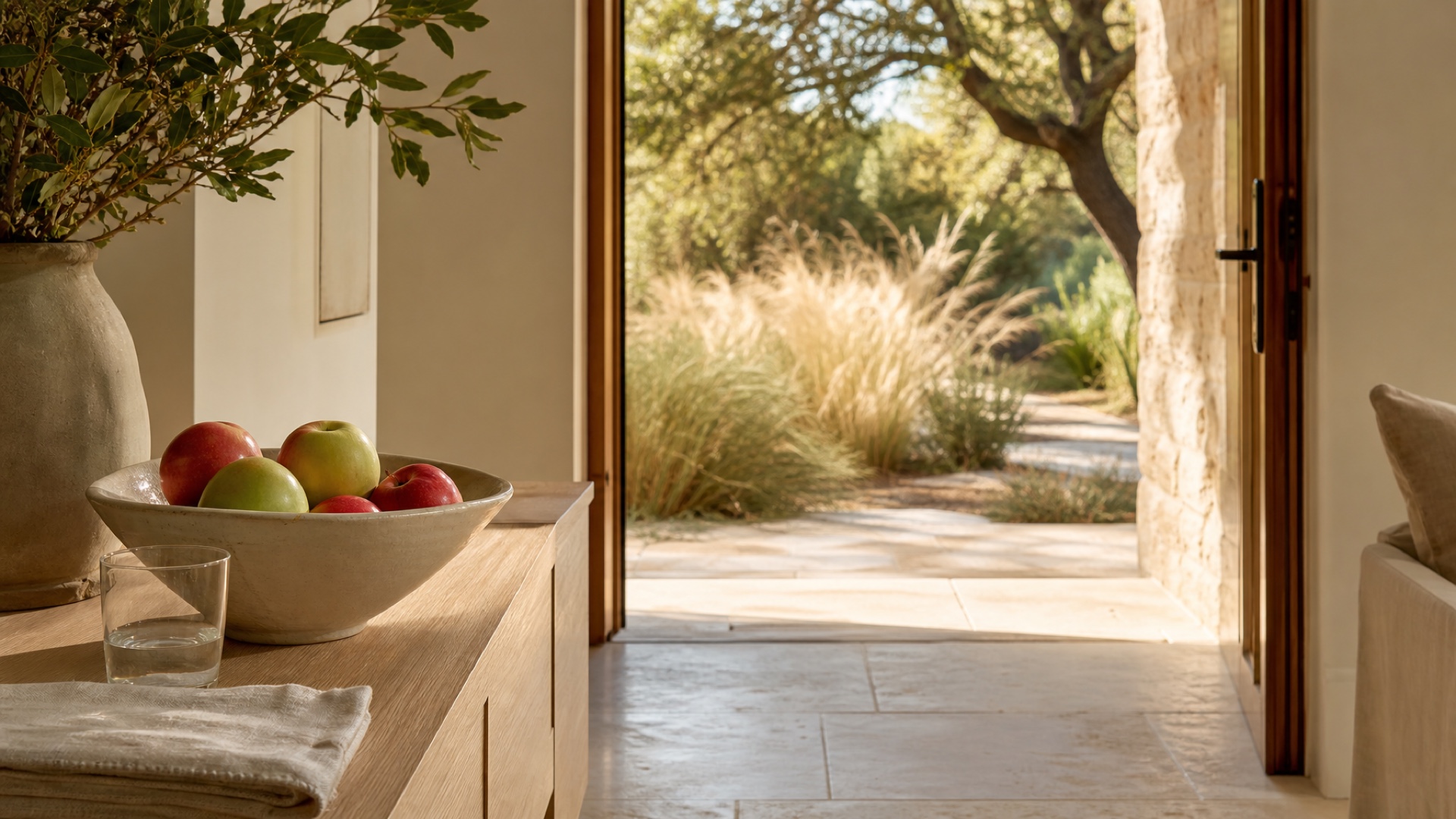 A bowl of apples and a glass of water on a pale-oak console beside an open doorway facing warm live-oak shade in late September.